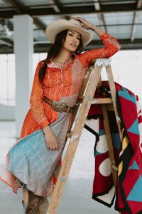 Ashley Callingbull leans on a ladder in a warehouse, wearing a cream cowgirl hat, a flowing orange dress, brown leather belt with large silver buckle.